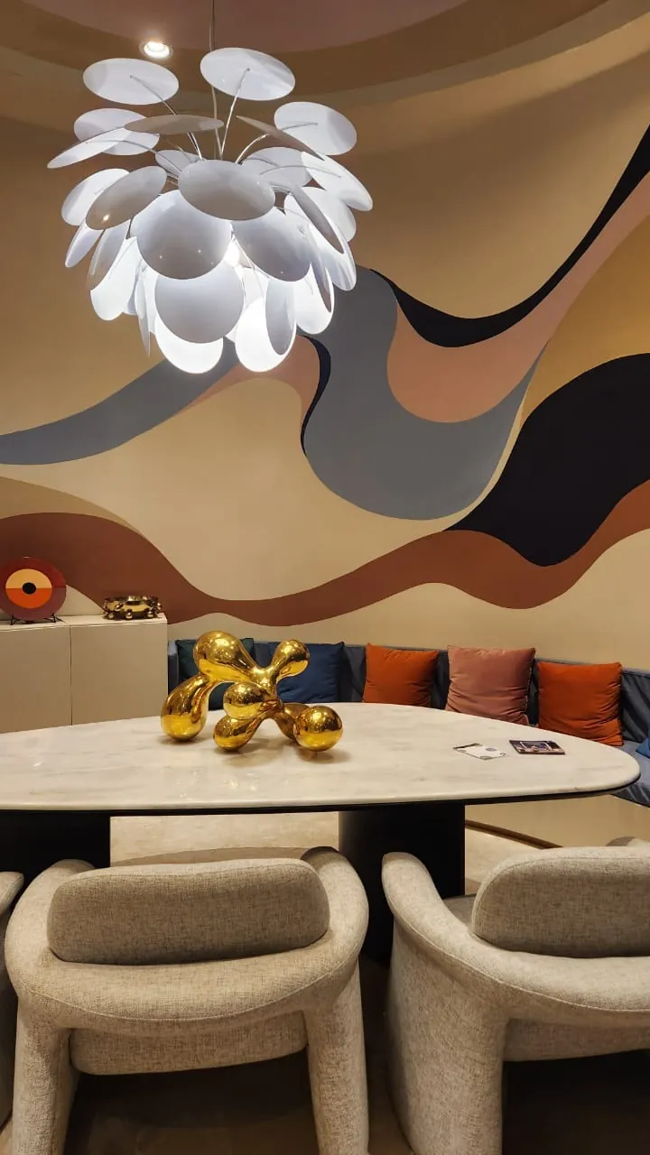 Maximalismo urbano: el color vuelve a ser protagonista (lo que vimos en Casa FOA 2025) Comedor con mural de ondas en tonos tierra, lámpara blanca de pétalos y sillas tapizadas