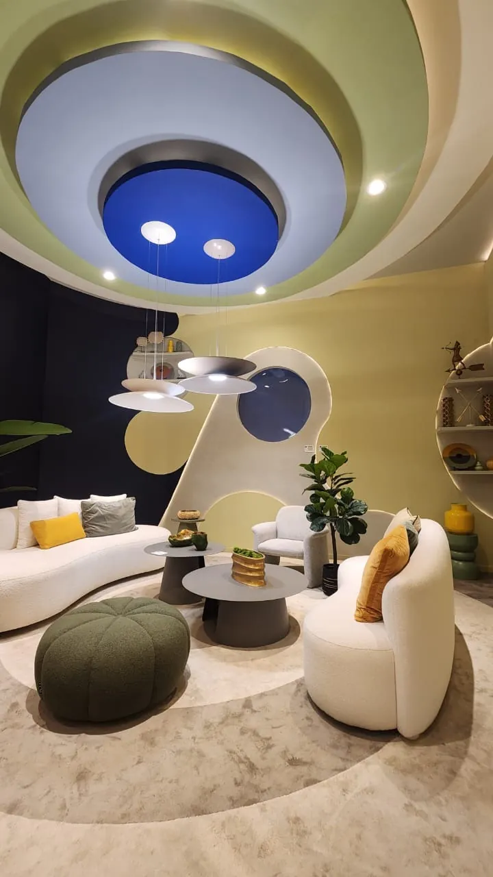 Maximalismo urbano: el color vuelve a ser protagonista (lo que vimos en Casa FOA 2025) Sala con techo en capas con círculo azul, sillones curvos blancos y puf verde